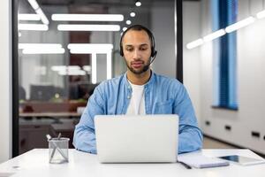 grave y enfocado joven Hispano hombre trabajando en estudiando a ordenador portátil en oficina vistiendo auriculares. foto