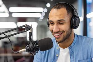 Close-up photo of a young smiling hispanic man in headphones sitting in front of a desk with a microphone in the office ,and talking online.