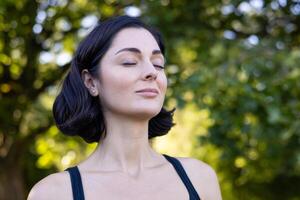 Close-up photo of a beautiful and athletic woman standing relaxed outside in the park and resting from training and running, closed her eyes and breathes deeply.