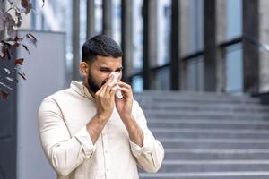 Close-up photo of a young Indian man standing near a building on the street and wiping his nose with a tissue, suffering from a severe runny nose and seasonal allergies.