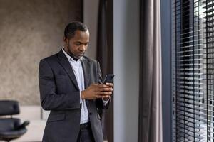 Concentrated black man in business suit holding cell phone in hands while standing next to window with jalousie. Attentive head of company solving business moments and making decisions at home. photo