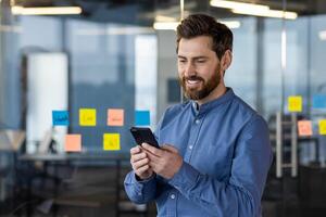 A young man is working and standing in the office against the background of a glass board with notes and smilingly using a mobile phone. photo