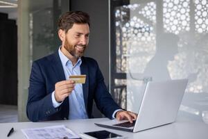 un sonriente y exitoso joven empresario en un traje se sienta en el oficina a un escritorio y trabajos en un computadora portátil, conduce en línea actas con un crédito tarjeta en su mano. foto