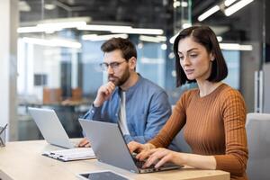 Caucasian woman sitting next to bearded man at shared workplace and typing on portable laptop. Ambitious colleagues working on personal results and solving list of tasks written on clipboard. photo