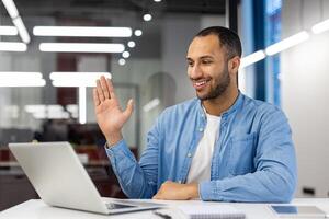 Cheerful Indian man at home in a modern living room engaged in a call, waving at the camera as if greeting a remote colleague or friend. photo