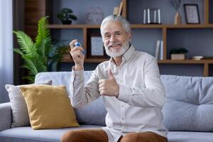 Cheerful elderly man sitting on a sofa, holding a hand grip strengthener and giving a thumbs up, showing positivity and active lifestyle in a cozy living room. photo
