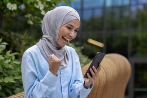 exitoso alegre musulmán mujer en hijab recibido en línea ganar notificación, dinka leyendo Noticias y participación mano arriba triunfo gesto celebrando sentado en banco fuera de oficina edificio en parque. foto