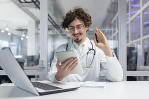 A man in a white medical coat with a stethoscope sits at a desk, engrossed in his work on a laptop and tablet. Doctor talking using call, greeting photo