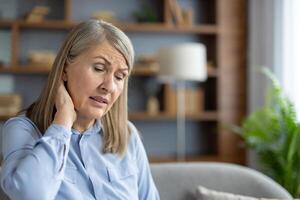 A senior Caucasian woman sitting in a living room appears distressed and uncomfortable, possibly experiencing neck pain or general malaise. photo