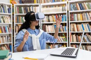 Young woman wears virtual reality headset in library, experiencing immersive technology with books and laptop around. photo
