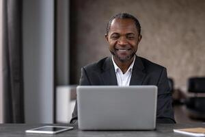 A professional African American man smiles while working on his laptop in an office setting. This image captures a moment of career satisfaction and confidence. photo