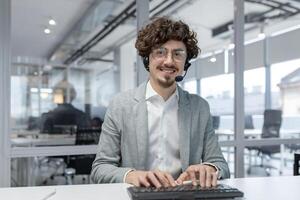 Portrait of a smiling young successful man wearing a headset working in the office at a computer, typing on a keyboard, talking to clients, looking confidently into the camera. photo