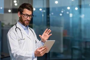 Joyful senior doctor in medical coat using tablet computer for call and online consultation with patient, man with beard and glasses in medical coat working inside clinic. photo