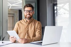 Portrait of a young businessman working in an office center, reading at a table with a laptop and holding a tablet in his hands, smiling and looking at the camera. photo