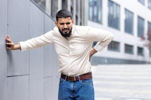 un cansado musulmán hombre es en pie fuera de propensión en contra el pared de el edificio, participación su mano detrás su atrás, haciendo muecas y sensación grave dolor en su inferior espalda foto