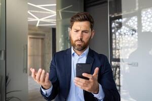 A worried young man businessman looks at the screen of the mobile phone he is holding in his hand and spreads his hands ,in surprise while in the office. photo