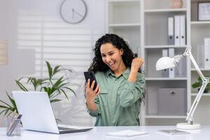 Happy hispanic woman sitting in the office at the table, rallying looking at the screen of the mobile phone, showing a victory gesture with her hand. photo