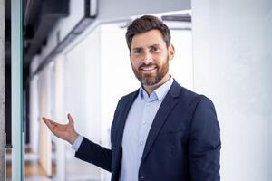 Portrait of a smiling, confident businessman in a modern office setting extending his hand as if presenting or greeting. photo