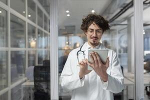 A doctor in a white lab coat and stethoscope holds a tablet in a medical office. photo