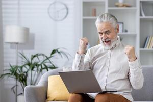 An elderly man with silver hair, comfortably sits on a sofa, jubilantly raising fists in triumph while looking at his laptop, symbolizing success or good news. photo