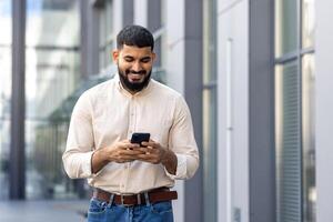 un sonriente joven musulmán hombre camina en el calle cerca negocio centros en un camisa y mira a el teléfono inteligente pantalla, textos, lee el Noticias foto