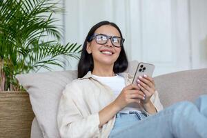 un joven Adolescente niña acostado en sofá a hogar y utilizando teléfono. recibe mensajes, chats con amigos foto