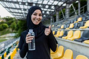 activo mujer en un hijab participación un agua botella, dando un pulgares arriba a un al aire libre estadio, demostración hidratación y salud. foto