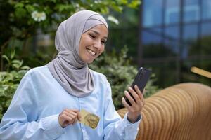 joven hermosa mujer en hijab con teléfono en manos y banco crédito tarjeta, musulmán fuera de oficina edificio reserva en línea y ordenando ropa, árabe mujer en en línea compras sonriente, satisfecho cliente. foto