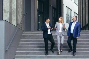 Interracial business meeting. Senior gray-haired man, young Asian man and one businesswoman in business suits walking up the stairs of a modern office center, talking to each other, discussing a deal. photo