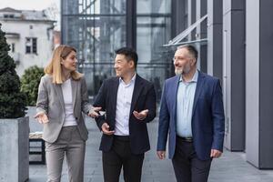 Diverse business group, three male and female workers walking and chatting discussing plans, outside office building photo