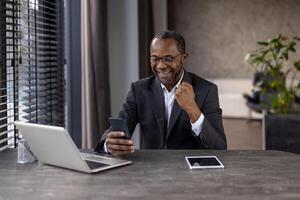 un sonriente africano empresario celebra éxito mientras utilizando su teléfono inteligente y ordenador portátil en un moderno oficina ajuste. foto