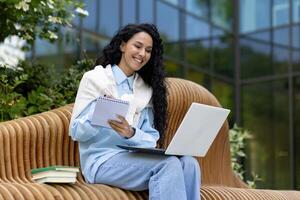 Young female student studying outside university campus, Latin American woman with laptop sitting on bench and recording class lecture, happy smiling. photo