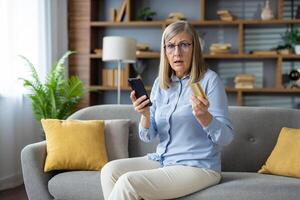 un maduro adulto mujer examina su crédito tarjeta mientras participación un teléfono inteligente, sentado cómodamente en su sofá a hogar, posiblemente suspicaz de fraude. foto