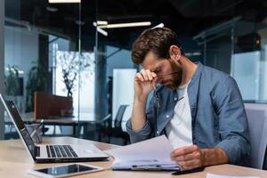 Upset businessman behind paper work inside modern office, mature man with beard reading financial reports and account documents unhappy with results and disappointed with achievements. photo
