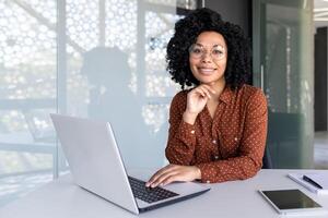 retrato de maduro adulto exitoso mujer de negocios, africano americano mujer a lugar de trabajo sonriente y mirando a cámara, confidente jefe trabajando a puesto de trabajo dentro oficina, utilizando ordenador portátil. foto