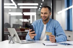 un persona atractivo en en línea compras utilizando un crédito tarjeta y teléfono inteligente a un contemporáneo oficina espacio de trabajo foto