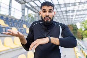 joven masculino atleta en ropa de deporte mira perplejo, gesticulando incertidumbre, mientras comprobación su reloj a un al aire libre estadio con asientos. foto