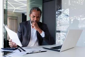 Serious thinking businessman behind paper work inside office, senior mature boss reviewing accounts contracts and financial reports, african american businessman solving accounting analytical tasks. photo