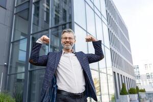 exitoso mayor empresario en traje en pie en frente de oficina edificio, levantamiento manos arriba, flexionando músculos, sonriente y mirando a cámara. foto