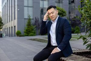 Tired young asian businessman in suit sitting on bench near working office and holding head, grimaced from pain and stress, closed eyes resting. photo