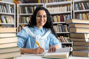 alegre Hispano hembra estudiante con lentes estudiando diligentemente entre imponente pilas de libros en un biblioteca. foto