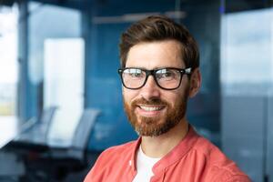 Close-up portrait of young businessman in red shirt, mature man in glasses smiling and looking at camera inside office near window, successful investor happy with achievements and good investment. photo