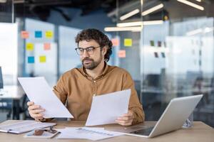 Thoughtful financial advisor in eyeglasses checking and analyzing printed graphs on paper sheets in personal workplace. Attentive man reading additional data for making deep research of investments. photo