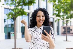 joven alegre mujer ganador recibido en línea notificación en teléfono, Hispano mujer con Rizado pelo celebrando éxito y triunfo caminando en ciudad cerca oficina edificio afuera. foto