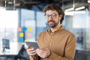 un alegre masculino profesional en un marrón camisa y lentes sostiene un digital tableta en un brillante, contemporáneo oficina espacio, exudando confianza y trabajo satisfacción. foto