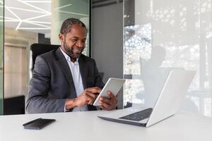 Mature adult african american boss investor at workplace using tablet computer, man happy with financial results and achievement smiling, using app inside office at table. photo