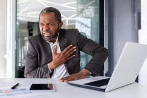 Heart attack in mature experienced man inside office at workplace, senior african american holding hands on chest, pain is severe in boss in business suit. photo