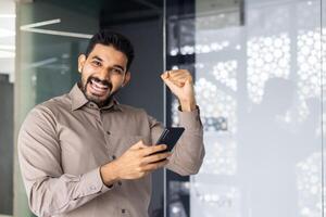 un alegre hombre en un casual camisa celebra éxito con un elevado puño en un oficina configuración, participación su teléfono. foto