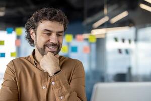 profesional masculino en un casual camisa trabajando y sonriente mientras mirando a el ordenador portátil en un brillante oficina ambiente con pegajoso notas en el antecedentes. foto