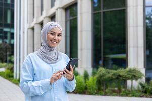 joven hermosa mujer de negocios en hijab camina en el ciudad desde fuera de el oficina edificio, mujer usos un solicitud en el teléfono, sonrisas y mira a el cámara, sostiene un teléfono inteligente para en línea llamar. foto